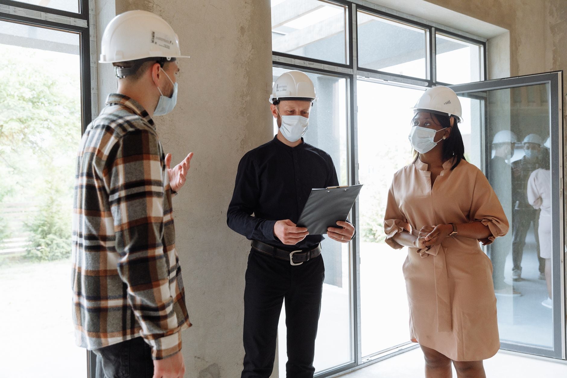 Real estate professionals discuss project indoors wearing masks and hard hats, ensuring safety.